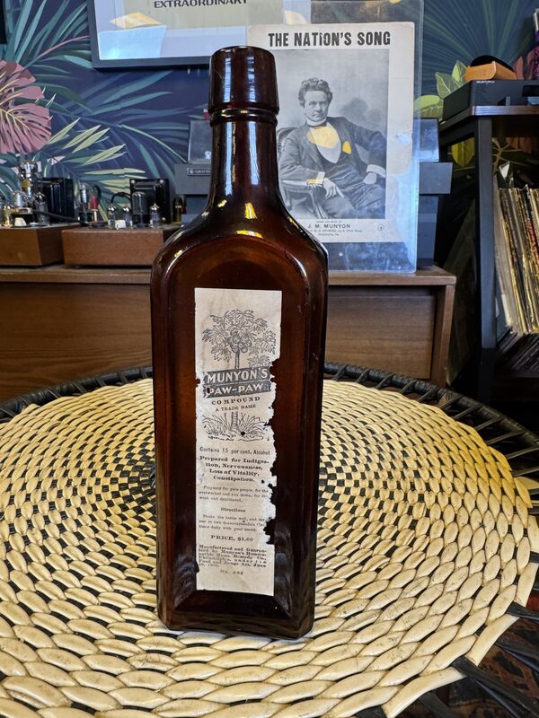 An amber rectangular glass bottle with a torn paper label reading 'Munyon's Paw-Paw Compound' stands centered on a round woven placemat. In the background are a wooden cabinet with audio equipment and vinyl records, and a framed black-and-white portrait titled 'THE NATION'S SONG.'