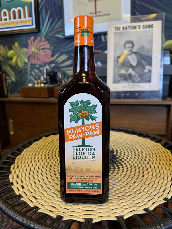 A tall brown glass bottle labeled 'Munyon's Paw-Paw Premium Florida Liqueur' with an orange cap stands centered on a round woven placemat on a table. Framed prints and decorative botanical-patterned wallpaper are visible blurred in the background.
