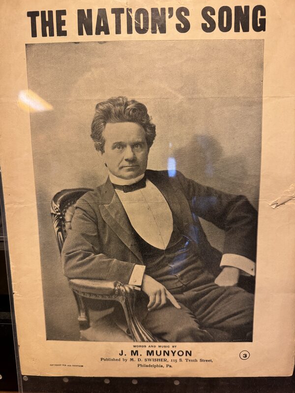 A vintage sheet-music cover titled 'THE NATION'S SONG' shows a black-and-white engraved portrait of a seated man in 19th-century formal attire reclining in an ornate chair. Text at the bottom reads 'J. M. MUNYON' with a Philadelphia publisher address, and the paper shows wear and glare reflections.