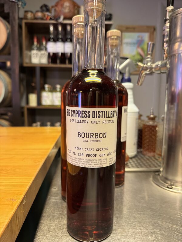 A tall glass bottle of dark bourbon with a white label reading "Big Cypress Distillery — Distillery Only Release — Bourbon — Cask Strength" (750 mL, 120 proof, 60% alc./vol.) stands on a metal bar counter in front of another similar bottle, with a wooden countertop to the left and shelves of bottles and barrels blurred in the background.