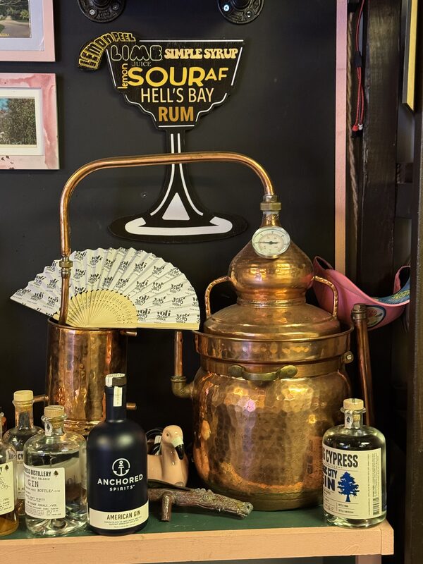 A hammered copper pot still with a domed lid, pressure gauge and connected copper tubing sits on a shelf against a black wall decorated with a cocktail-glass-shaped sign reading "SOUR AF HELL'S BAY RUM."