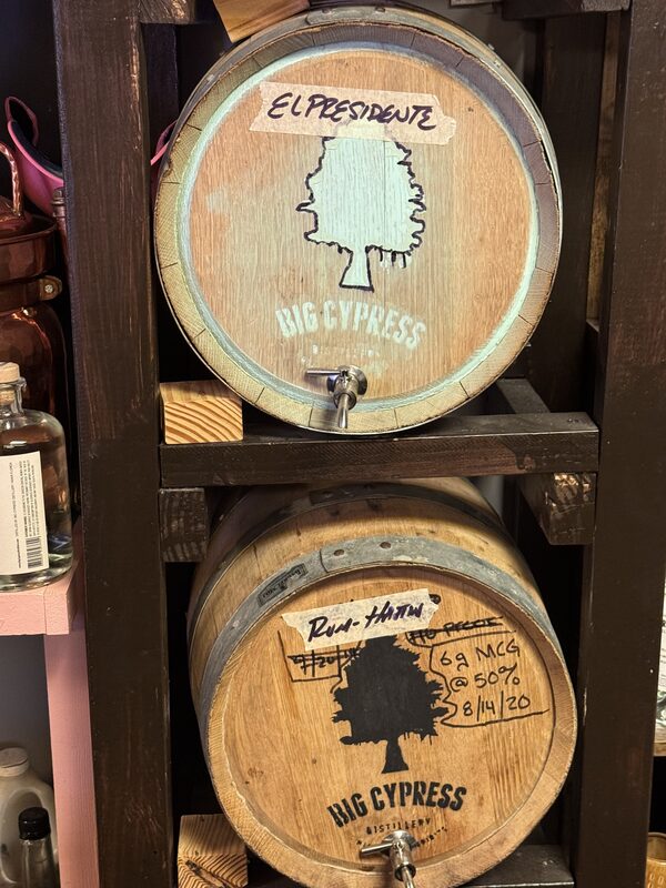 Two wooden barrels stacked in a dark wooden rack, each stamped "BIG CYPRESS" with a tree silhouette; the top barrel has a taped handwritten label reading "EL PRESIDENTE."
