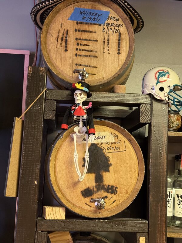 Two wooden barrels stacked in a dark wooden rack, the top barrel bearing a blue tape label reading "WHISKEY 2/19/24"; a small skeleton figurine dressed in a hat and suit sits on the edge of the lower barrel.