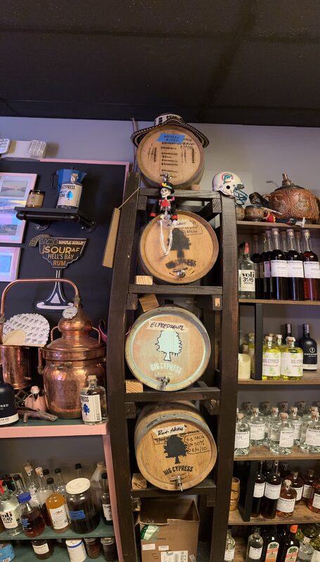 A tall dark wooden rack holds four small labeled oak barrels, flanked by shelves and countertops filled with bottles of liquor, a copper still, and a painted skull.