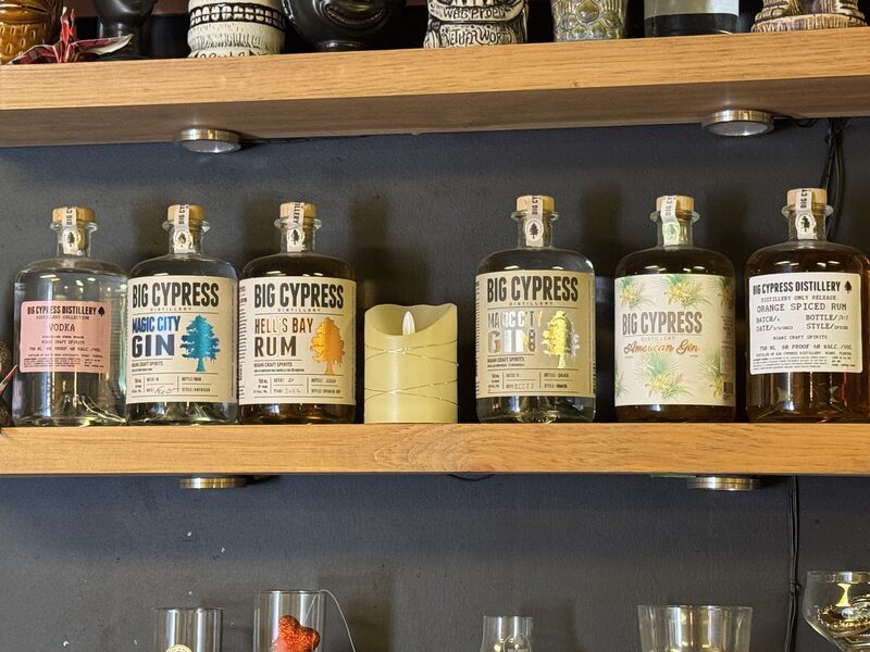 A wooden shelf displays seven clear glass bottles of Big Cypress Distillery spirits, including vodka, Magic City gin, Hell's Bay rum, American gin, and orange spiced rum, arranged side by side.