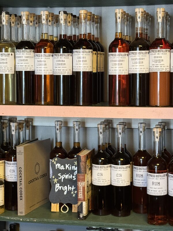 Two shelves filled with rows of tall glass bottles holding variously colored liqueurs and spirits, with cocktail books including "Cocktail Codex" on the lower shelf.