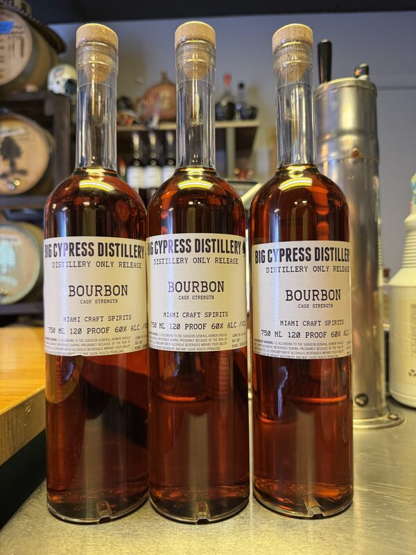 Three clear glass bottles of Big Cypress Distillery Bourbon with cork stoppers and white labels reading "Bourbon Cask Strength" stand side by side on a metal counter, each filled with amber liquid.