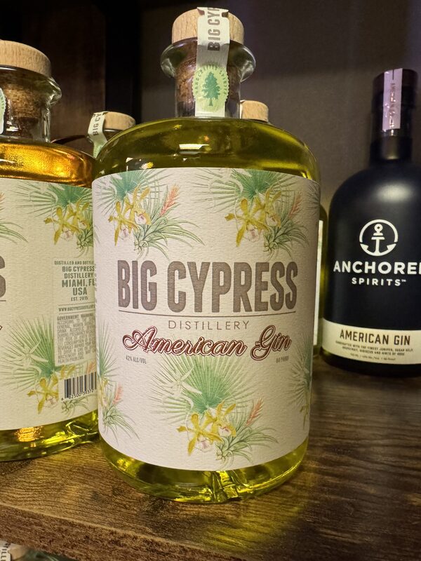 A clear glass bottle of Big Cypress Distillery American Gin with a floral label and cork stopper sits on a wooden shelf, containing a pale yellow liquid, next to a black bottle labeled "Anchored Spirits American Gin."