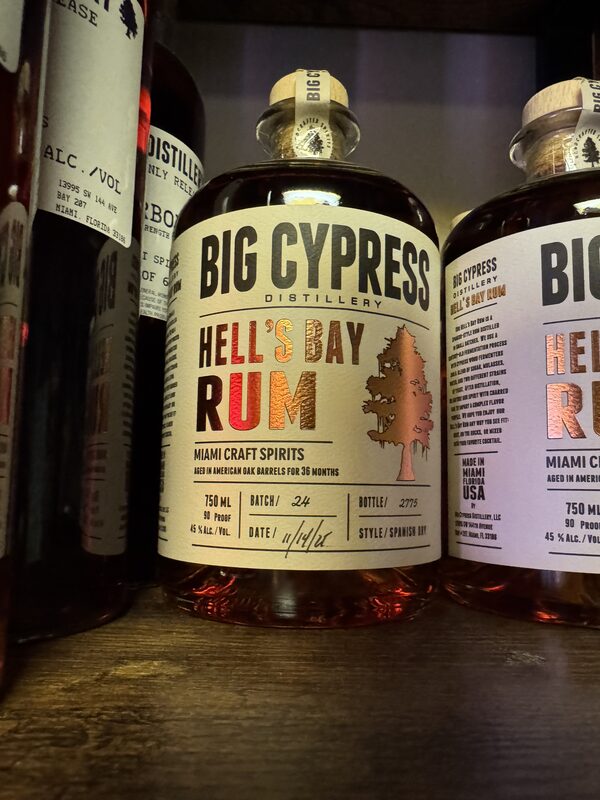 A rounded glass bottle of amber rum centered on a wooden shelf with a cream label reading "BIG CYPRESS" and "HELL'S BAY RUM" in metallic red and gold, featuring a stylized tree silhouette.