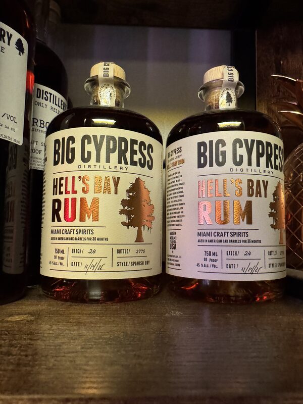 Two squat glass bottles of Big Cypress Distillery "Hell's Bay Rum" sit side by side on a wooden shelf, each with a cork stopper and cream label featuring a copper-toned tree silhouette.