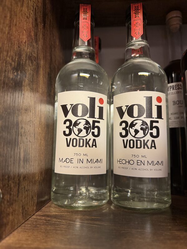 Two clear glass bottles of "Voli 305 Vodka" with white labels and globe logos sit side by side on a wooden shelf; one label reads "MADE IN MIAMI" and the other "HECHO EN MIAMI."