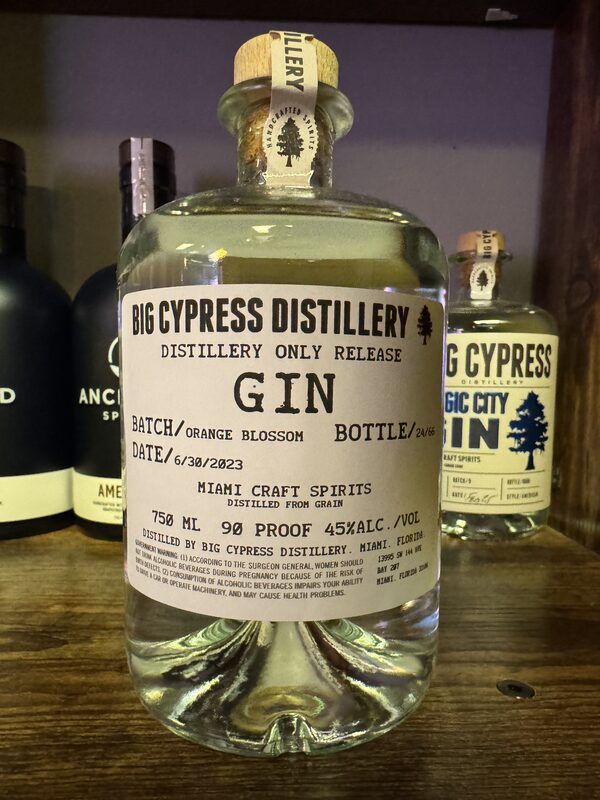 A clear glass bottle of Big Cypress Distillery gin with a cork stopper and a large white label reading "GIN" (batch "Orange Blossom") sits centered on a wooden shelf.