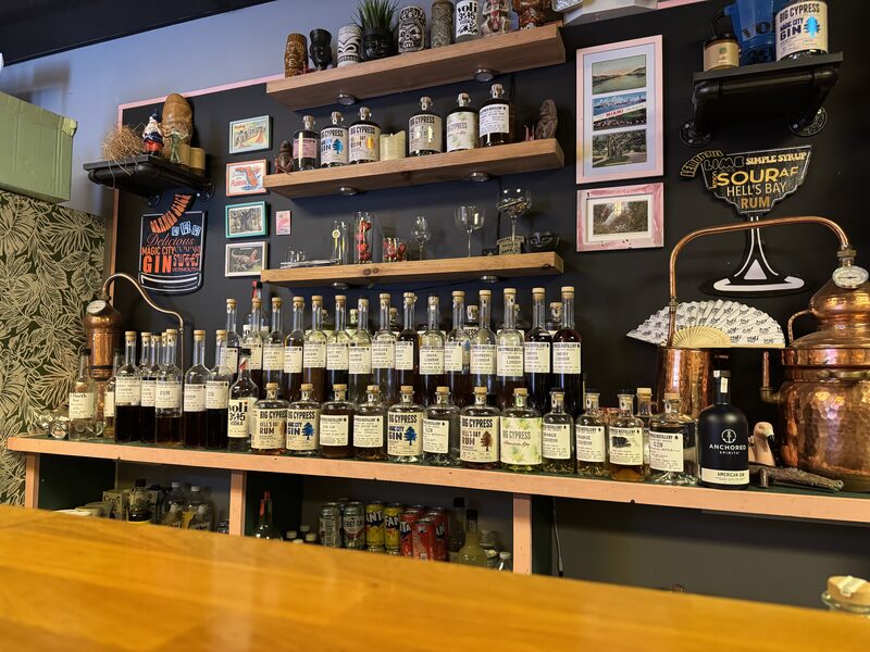 A bar display showing multiple rows of labeled bottles arranged on a countertop and wooden wall shelves, with glassware, tiki mugs, and branded signs on a dark wall behind.