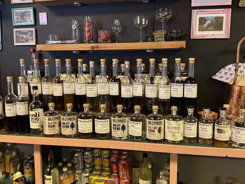 A dark-walled shelving display holds two wooden shelves and a countertop lined with clear-glass liquor bottles with cork stoppers and white labels reading "Big Cypress," with a copper distilling pot at the right.
