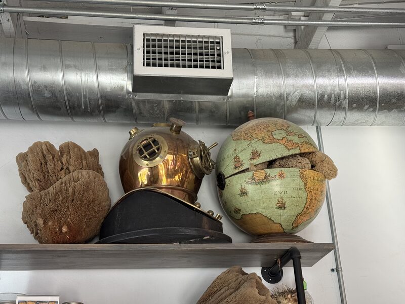 A wooden shelf mounted against a white wall beneath an exposed metal air duct holds a brass antique diving helmet centered between two large natural sea sponges. To the right of the helmet is a decorative globe with a large broken section revealing its interior.
