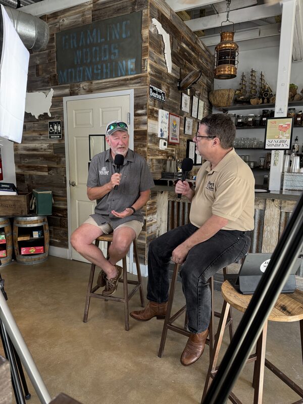 Two men sit on wooden stools holding handheld microphones in a rustic tasting-room or bar with weathered wood walls, shelves of glassware and bottles, a hanging lantern and a model ship behind them. One man wears a gray shirt, shorts and a baseball cap with sunglasses on the brim, and the other wears a beige polo, jeans and brown boots.