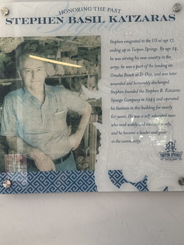 A mounted decorative plaque showing a sepia-toned portrait of an older man standing with one hand on his hip at left and a block of printed biographical text at right beneath the heading "HONORING THE PAST." The plaque features a patterned blue border along the bottom, a small circular logo at lower right, four visible metal mounting screws in the corners, and reflections/glare across the photograph.