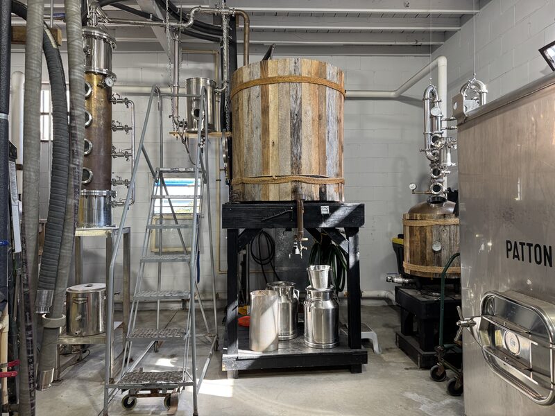 An indoor distillery room with a large wooden fermentation barrel mounted on a black stand at center and stainless steel jugs and a funnel beneath it. The barrel is flanked by tall stainless steel column stills, piping and a wheeled metal ladder on the left, and a stainless steel unit labeled "PATTON" on the right.