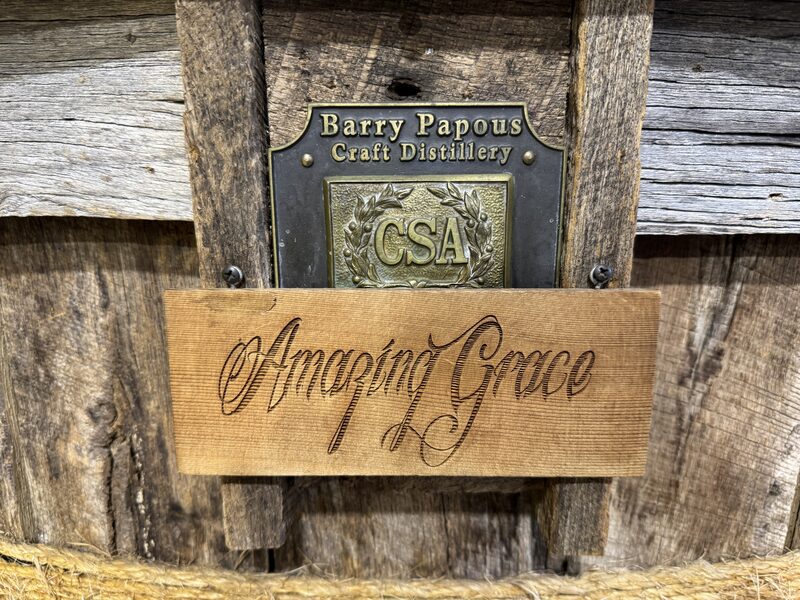 A weathered wooden barrel or plank surface with a metal plaque mounted near the top that reads "Barry Papous Craft Distillery" above a decorative "CSA" emblem. A rectangular wooden tag engraved in flowing script with the words "Amazing Grace" is attached below the plaque.