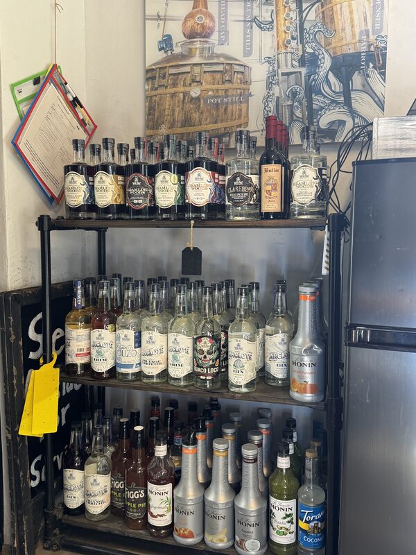 A three-tier black metal shelving unit filled with rows of liquor and syrup bottles—clear and dark spirits, labeled bottles with paper tags, and several silver Monin syrup bottles—occupies the center of the image. Behind the shelves is a large distillery-themed poster on the wall and clipboards hanging to the left, with a refrigerator visible on the right.